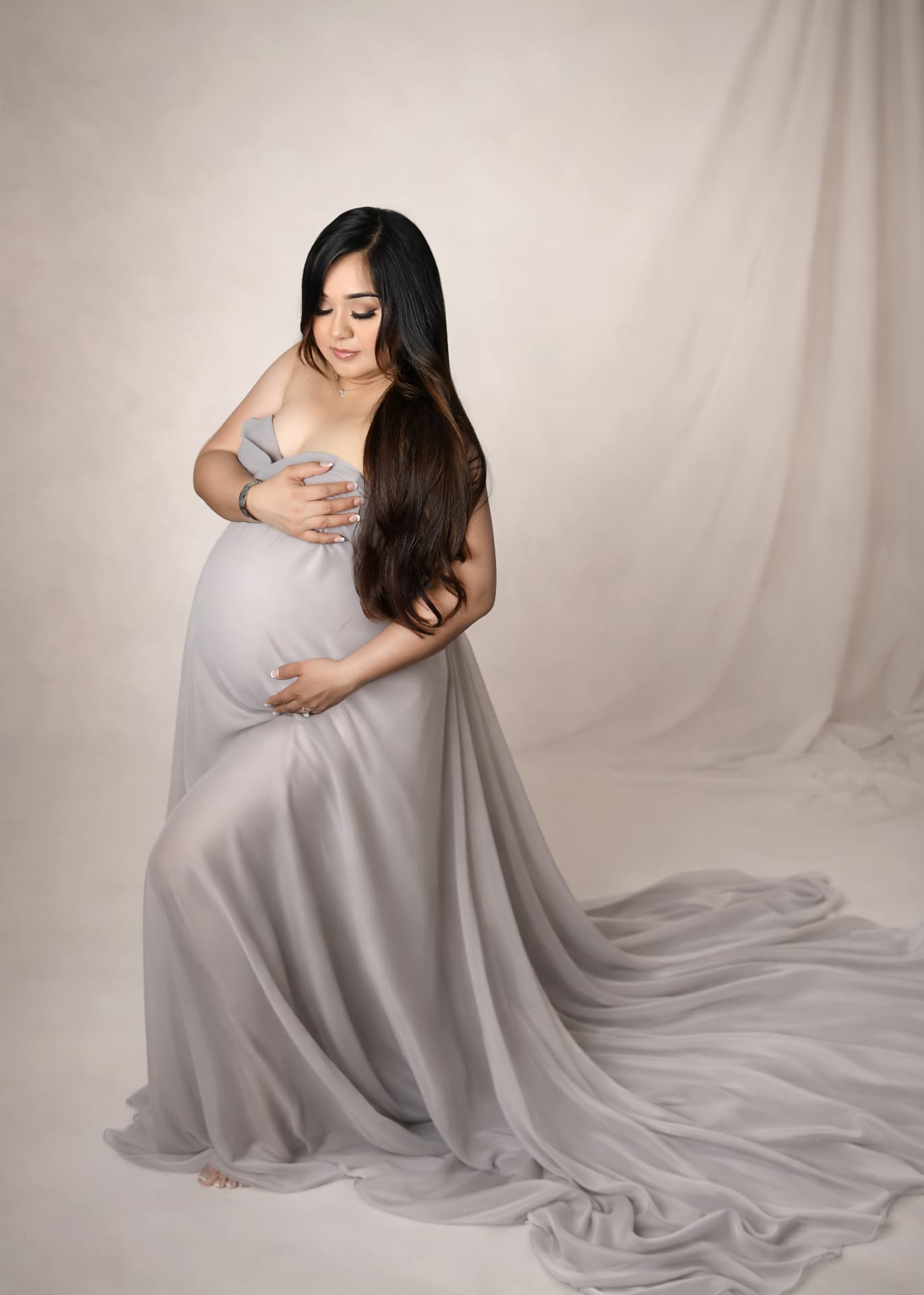 Studio maternity portrait in a flowing grey gown on a cream backdrop