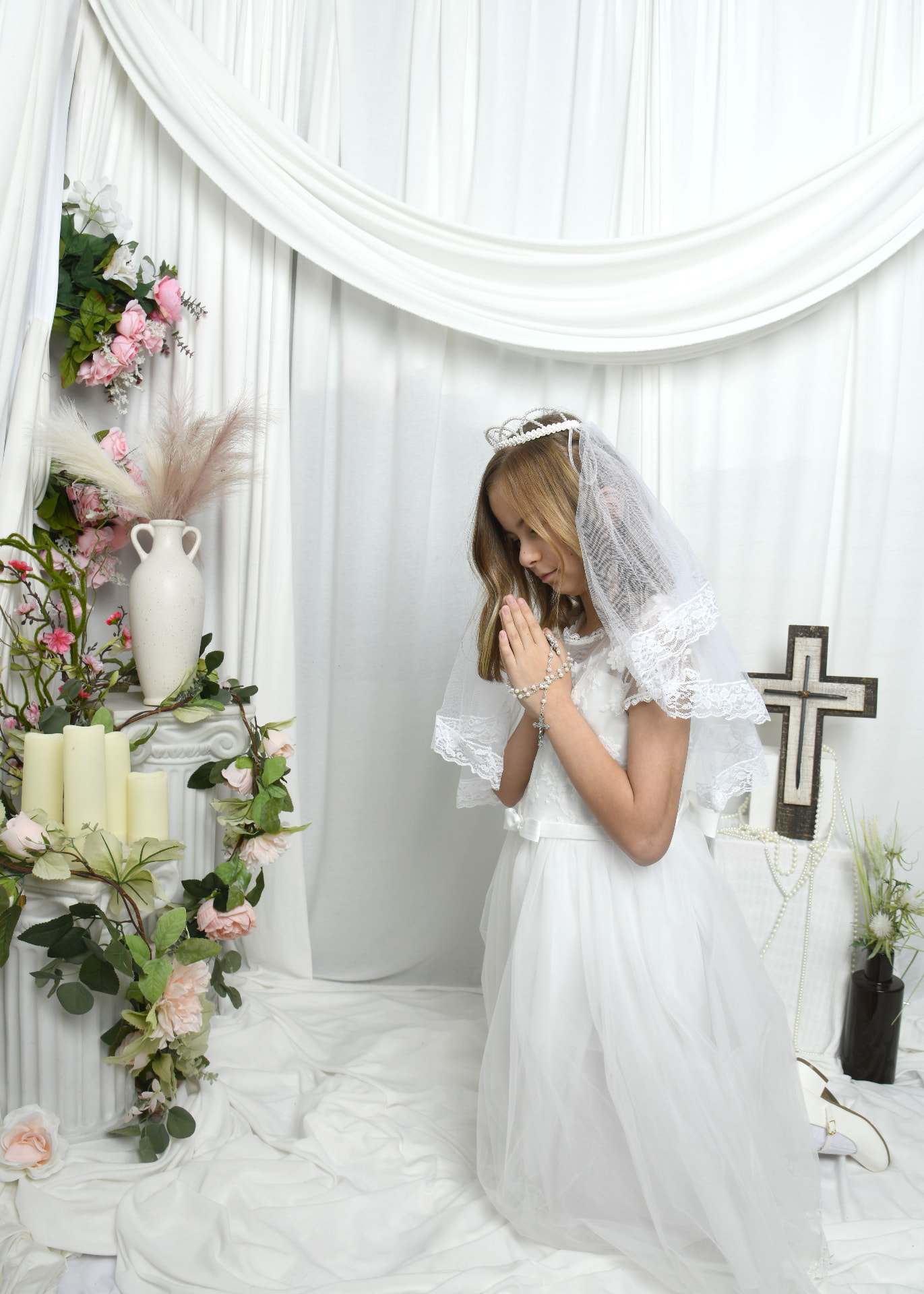 Girl in a white First Communion gown praying with a rosary beside candles and a cross